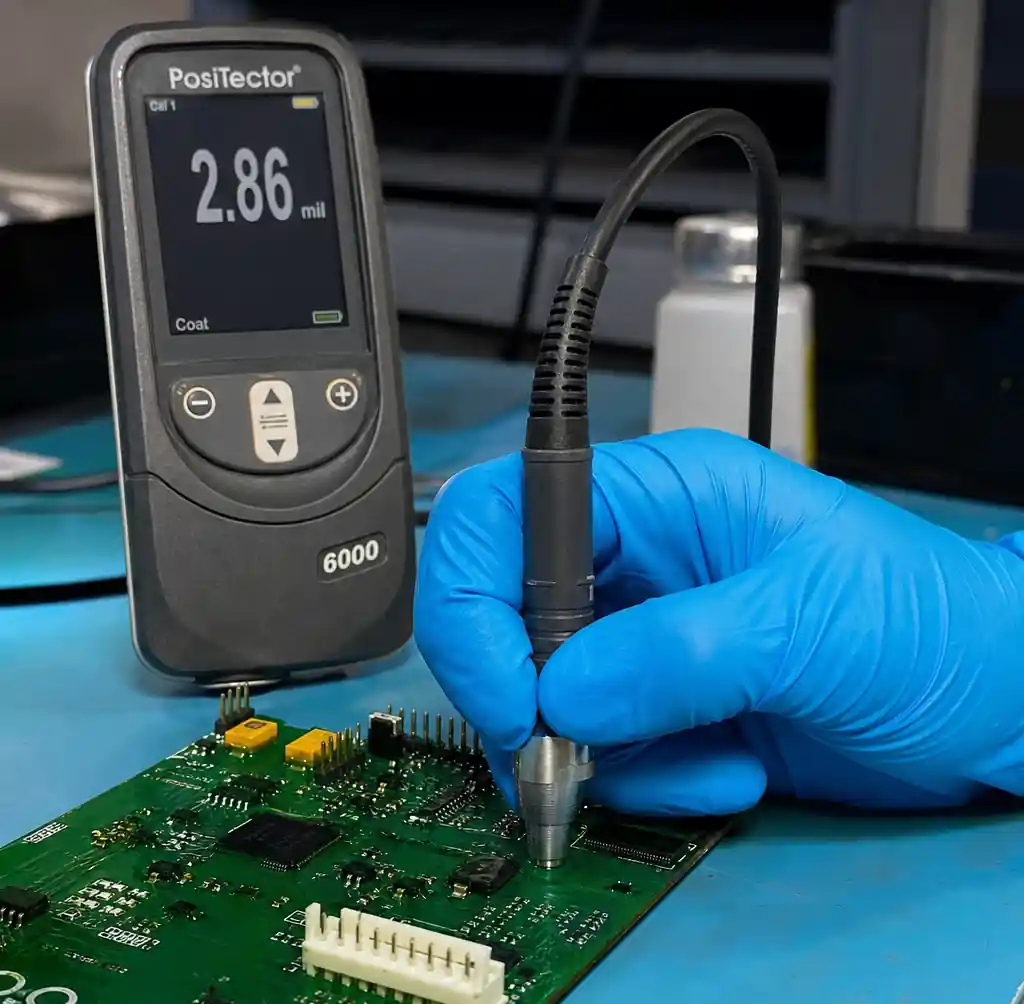 Technician measuring conformal coating thickness on a PCB with a wet film or eddy-current gauge